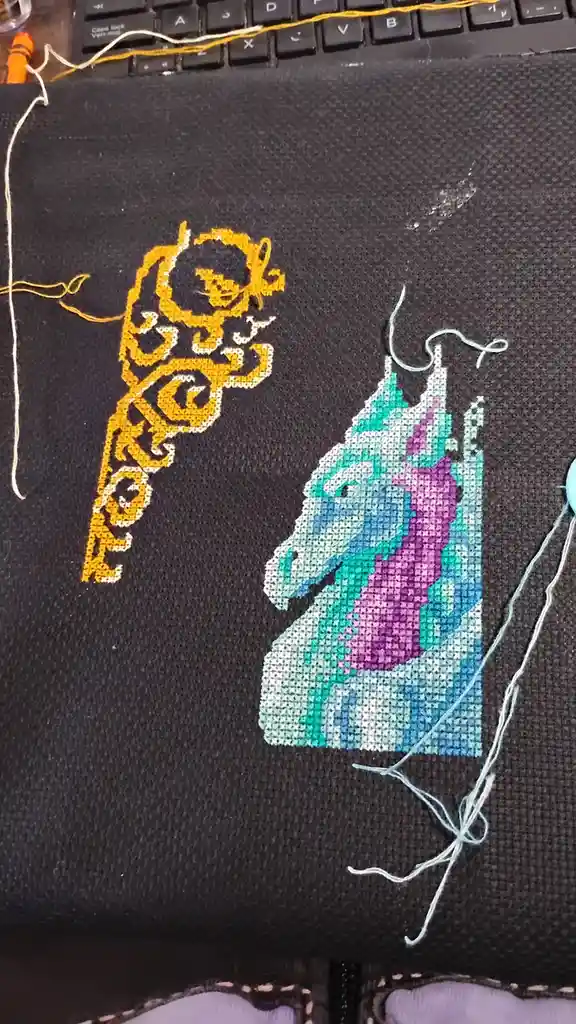 A close-up photo shows two partially completed cross-stitch designs on dark fabric: a yellow ornate pattern resembling a…