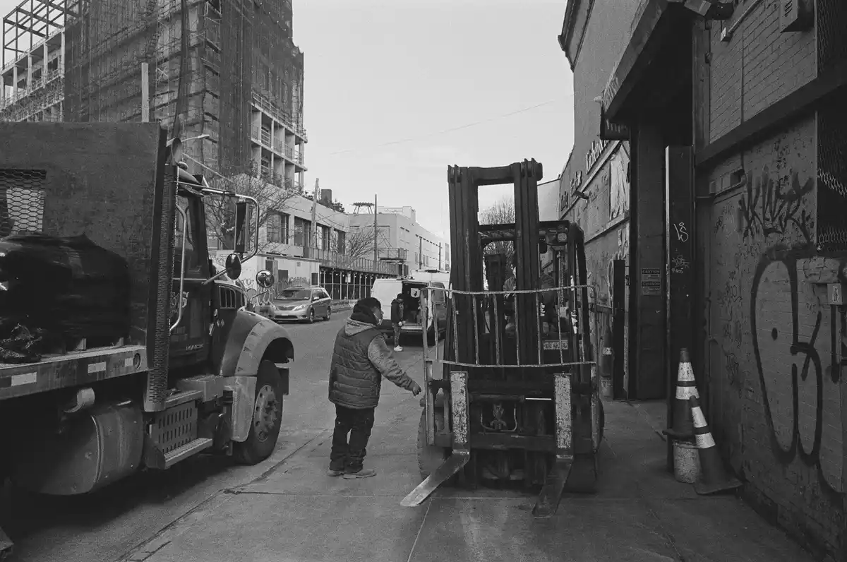 A black-and-white photo shows a man in a puffy vest standing on a sidewalk between a large truck and a forklift, gesturing…