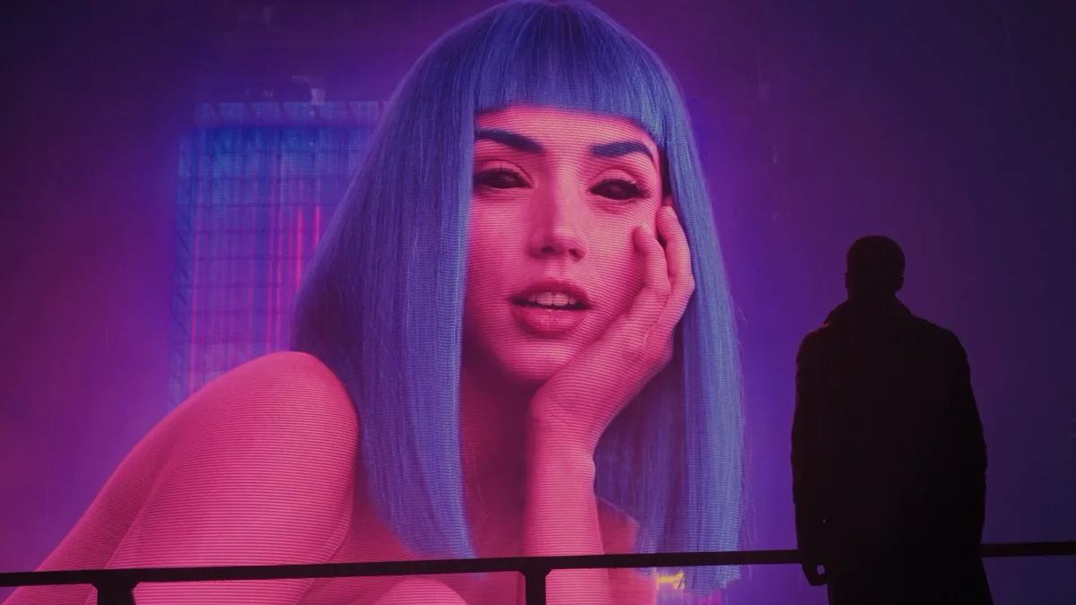 A person stands in silhouette, looking at a large, glowing digital billboard displaying a woman with blue hair and a hand on…