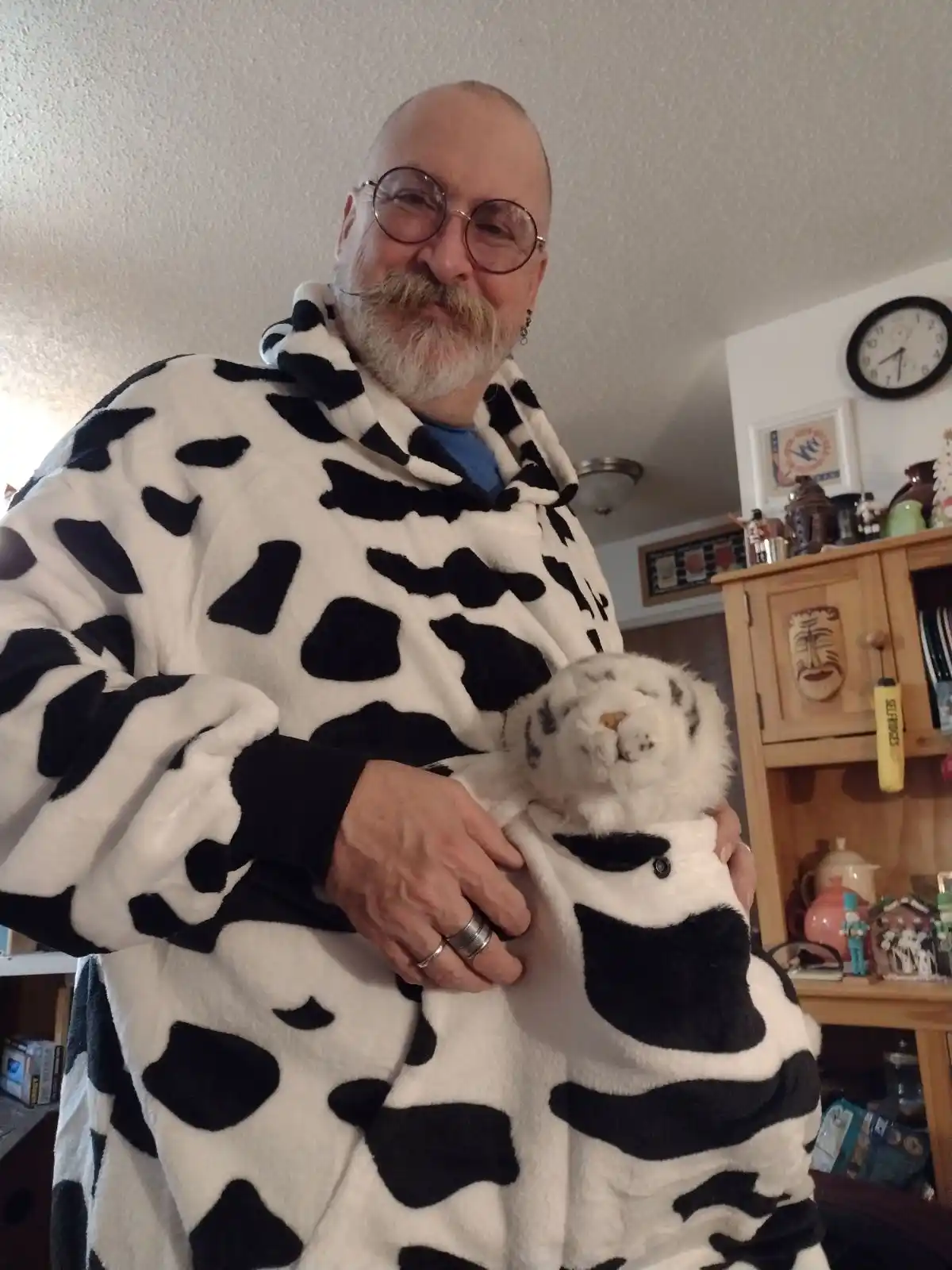 A man with glasses and a mustache wears a black-and-white cow-print robe and holds a stuffed white tiger. The text overlaid…
