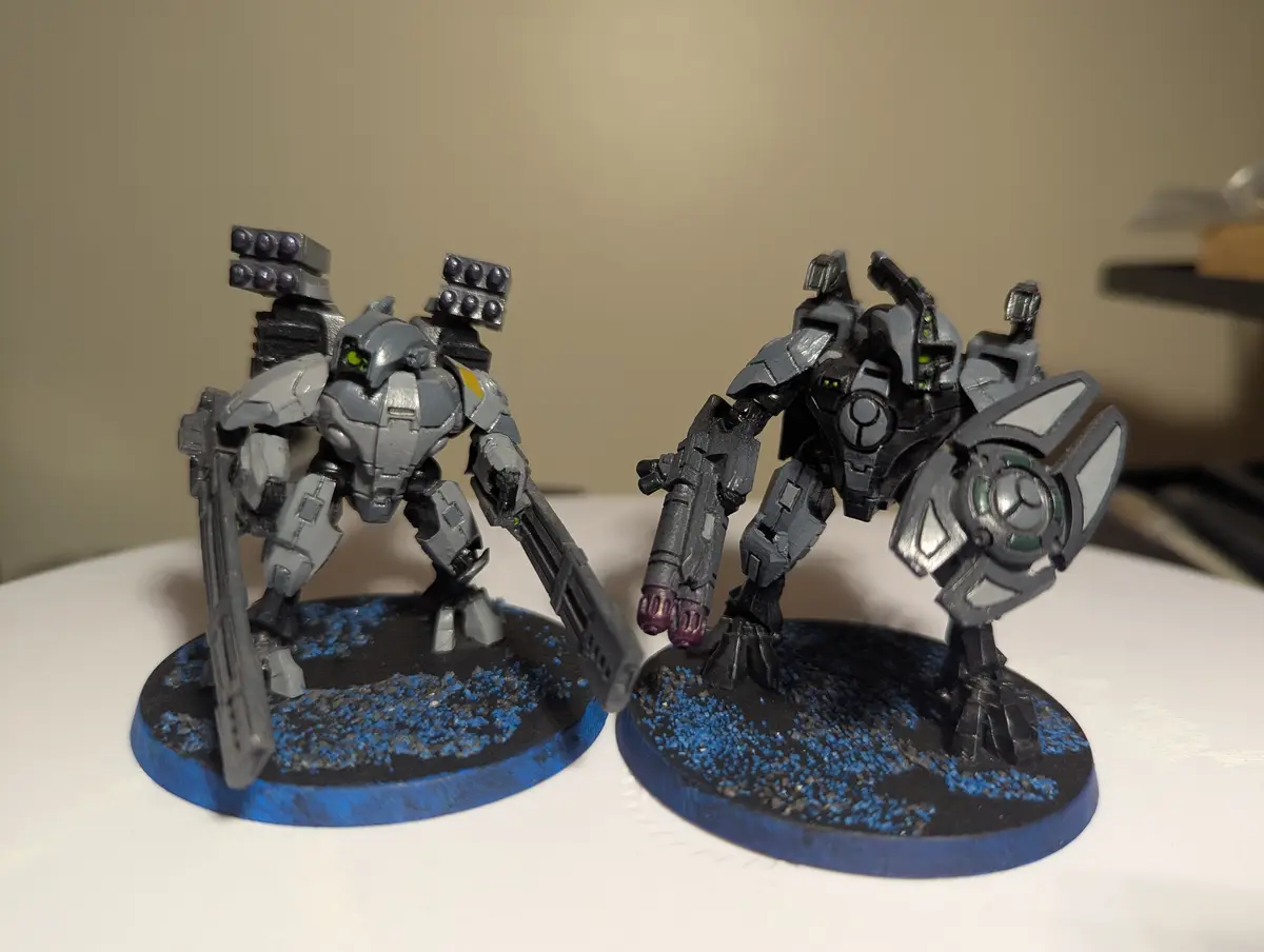 Two painted miniature mechs stand side-by-side on circular bases with blue speckled terrain, both featuring glowing green…