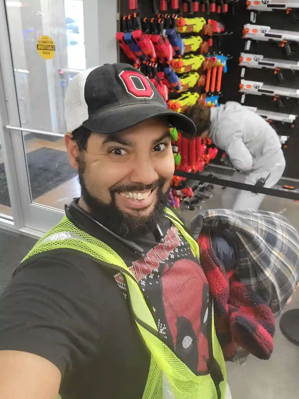 A man in a bright yellow safety vest and a black-and-white baseball cap with a red 'O' smiles at the camera while holding a…