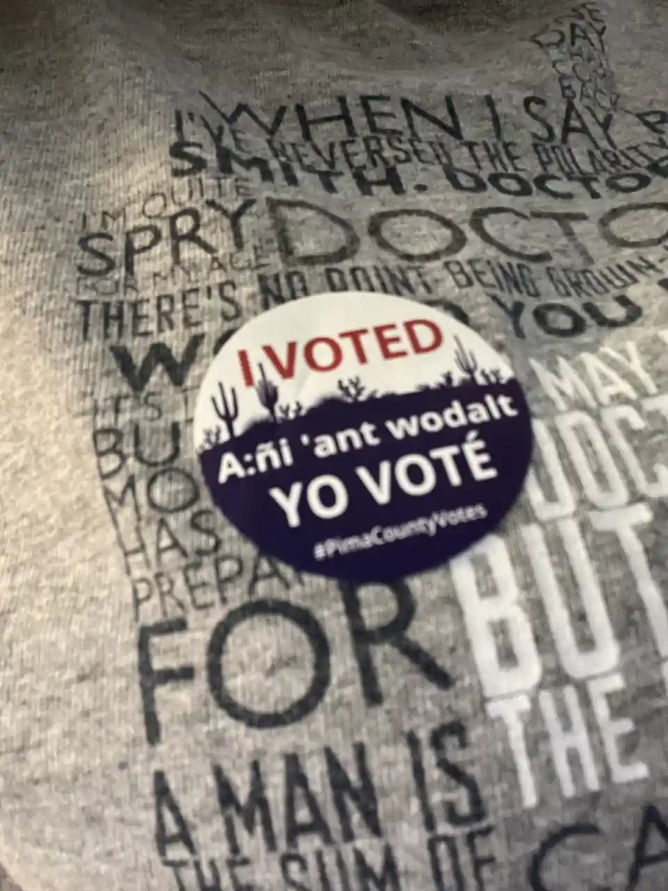 A close-up of a circular 'I VOTED' sticker on a gray t-shirt with faded text. The sticker has 'I VOTED' in red at the top, a…