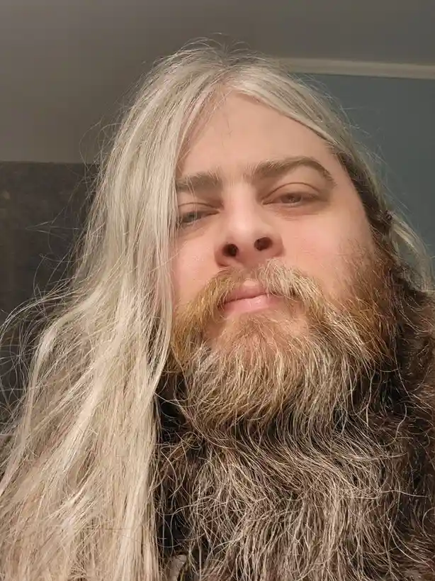 A close-up selfie of a person with long, straight, platinum blonde hair and a full, long beard with mixed brown and gray…