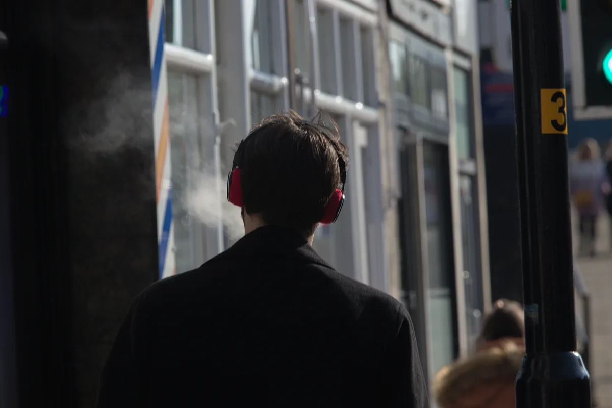 A person viewed from behind walks down a city street wearing red and black over-ear headphones. A visible plume of white…