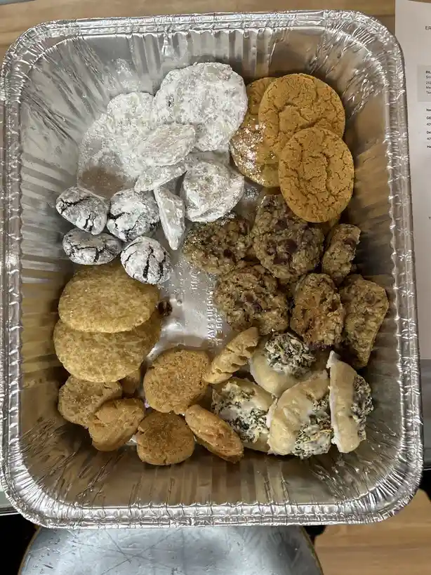 A top-down photo of a large aluminum baking tray filled with various types of cookies, including powdered sugar cookies…