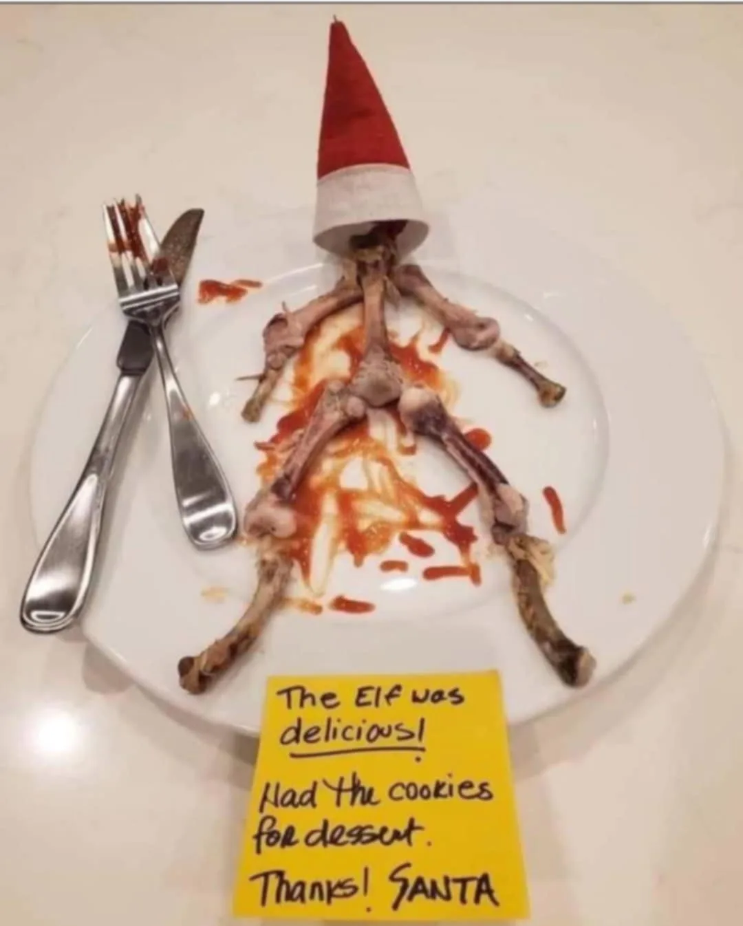 A white plate holds leftover chicken bones arranged to resemble a human figure wearing a red Santa hat, with sauce stains…