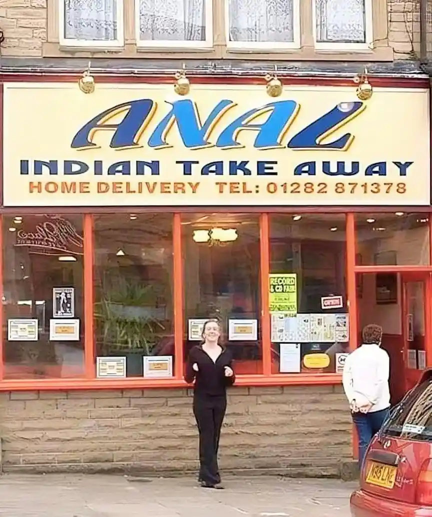 A woman stands smiling in front of a restaurant with a large sign that reads 'ANAL INDIAN TAKE AWAY' with 'HOME DELIVERY…