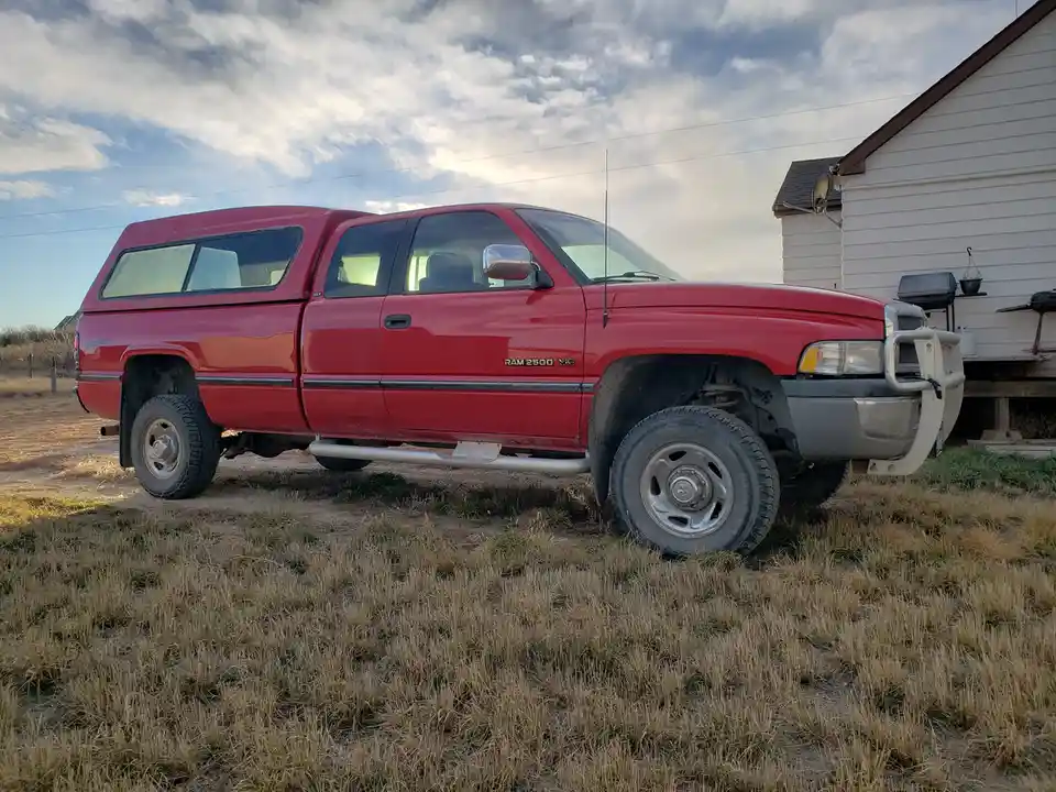 A red Dodge Ram 2500 pickup truck with a camper shell is parked on dry grass in front of a white house under a cloudy sky…