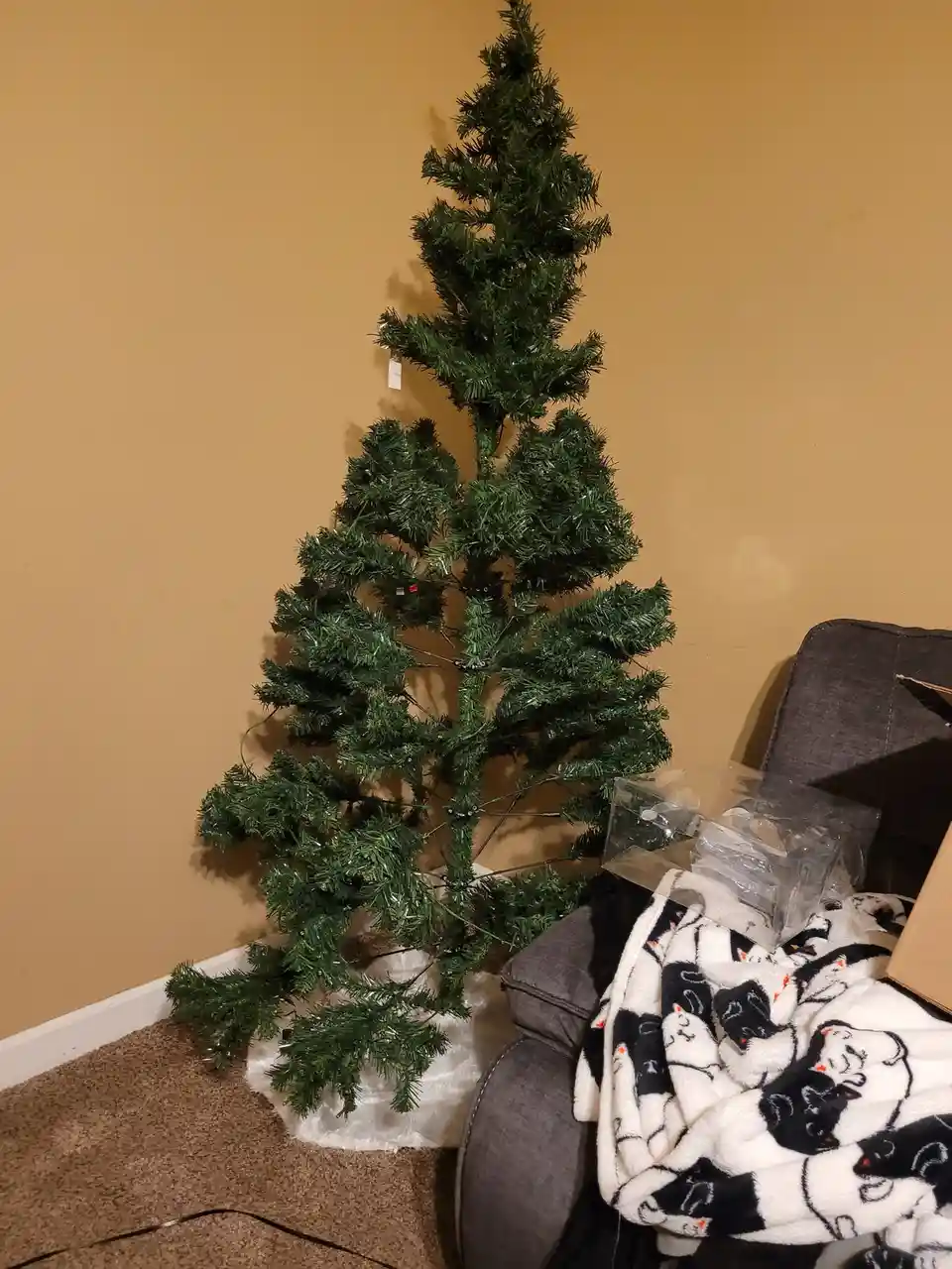 An unadorned, artificial Christmas tree stands in a corner of a room with beige walls and brown carpet, next to a gray…