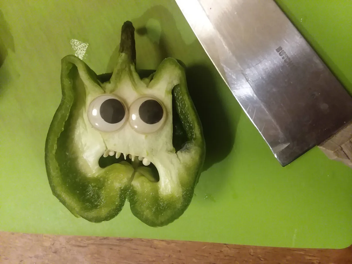 A halved green bell pepper on a green cutting board has googly eyes glued to its top and its inner seeds arranged to look…