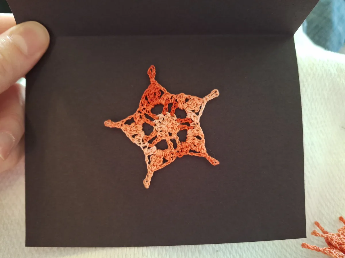 A hand holds open a black card revealing a crocheted star-shaped ornament in shades of orange and peach, with visible yarn…