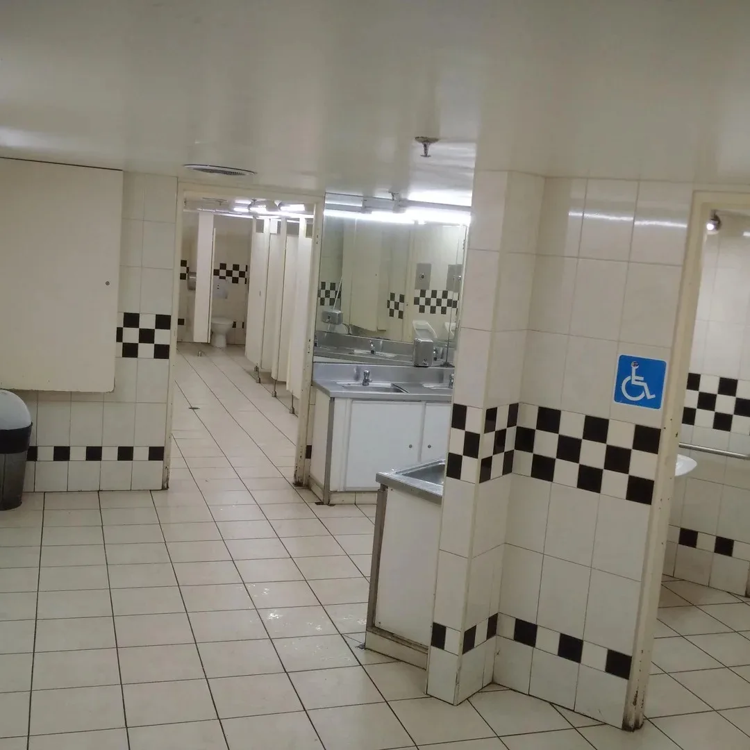 a photo of a public restroom with white tiled floors and walls, featuring a blue wheelchair-accessible symbol on a stall…