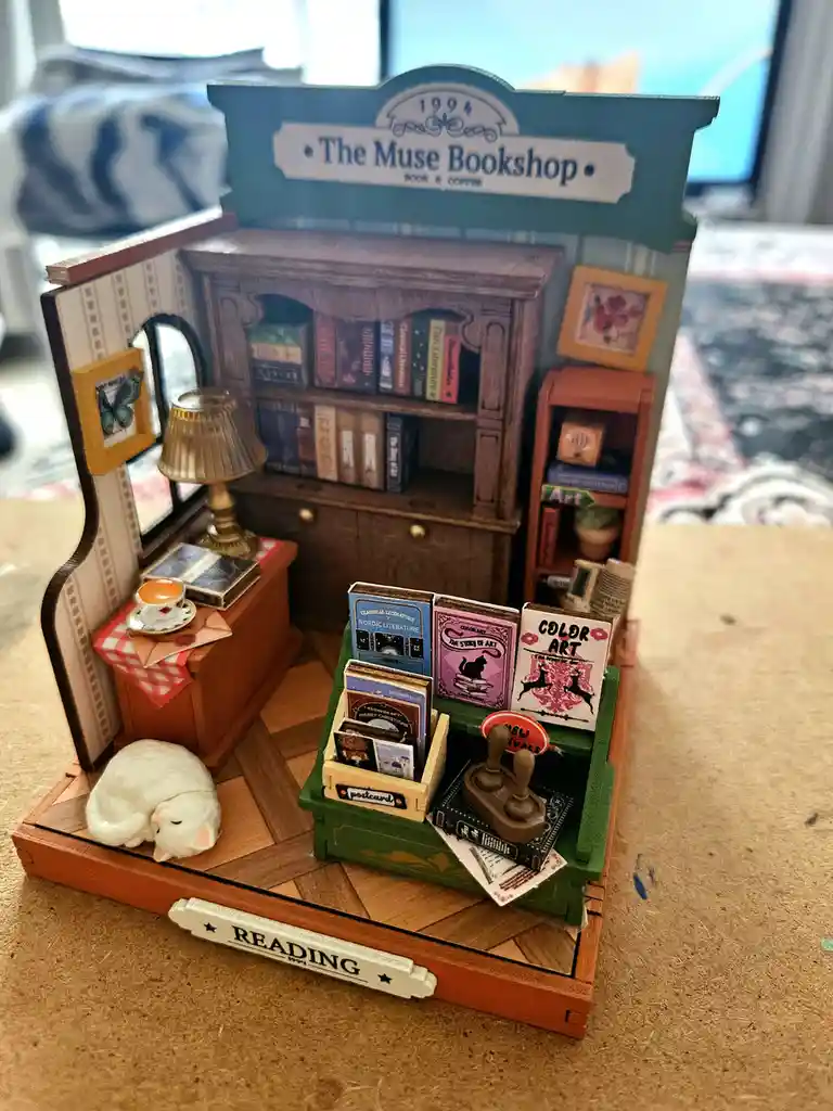 A miniature diorama of a cozy bookshop named 'The Muse Bookshop' from 1994, featuring tiny bookshelves, a lamp, a cup of…