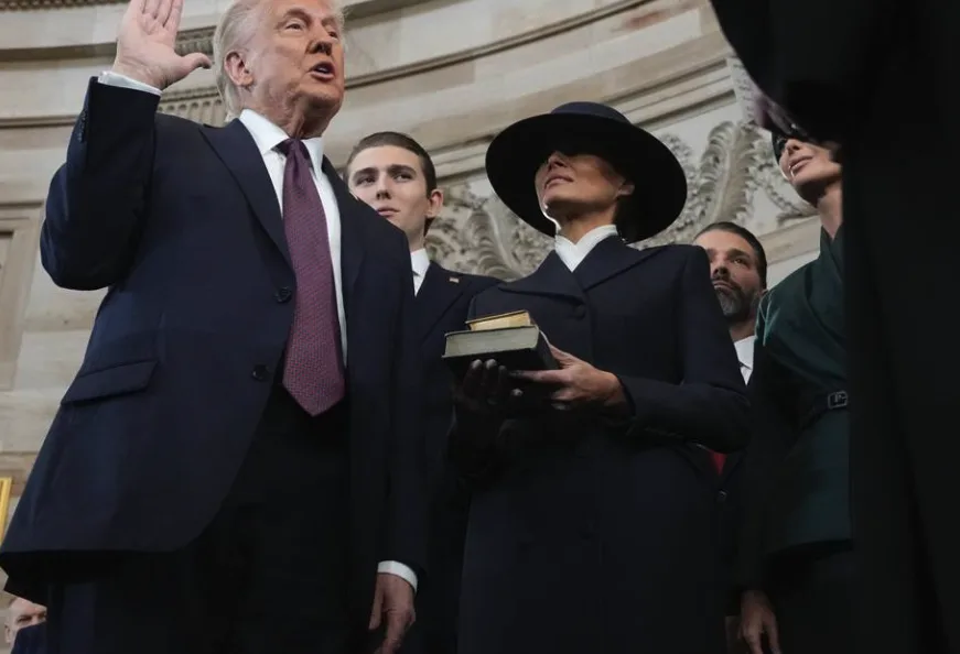 A man in a dark suit and tie raises his right hand while swearing an oath, standing beside a woman in a dark coat and…