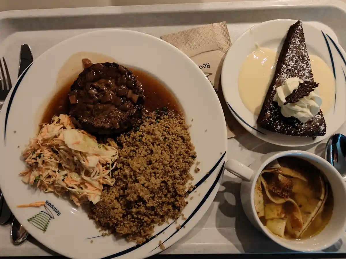 A meal tray contains a large steak with sauce, quinoa, and coleslaw on a plate, alongside a slice of chocolate cake with…