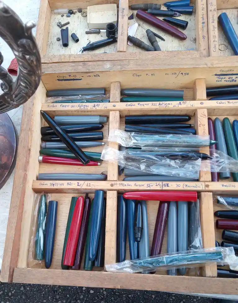 A wooden display case filled with neatly organized vintage fountain pen nibs, barrels, and clips, labeled with handwritten…