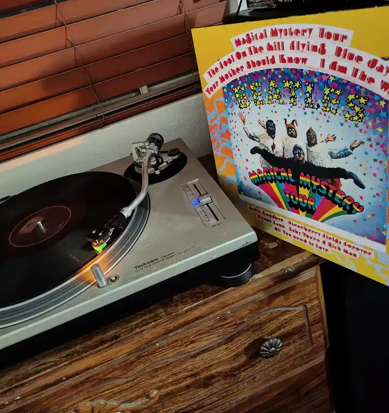 A Technics turntable sits on a wooden dresser next to a Beatles 'Magical Mystery Tour' album cover. The album art features…