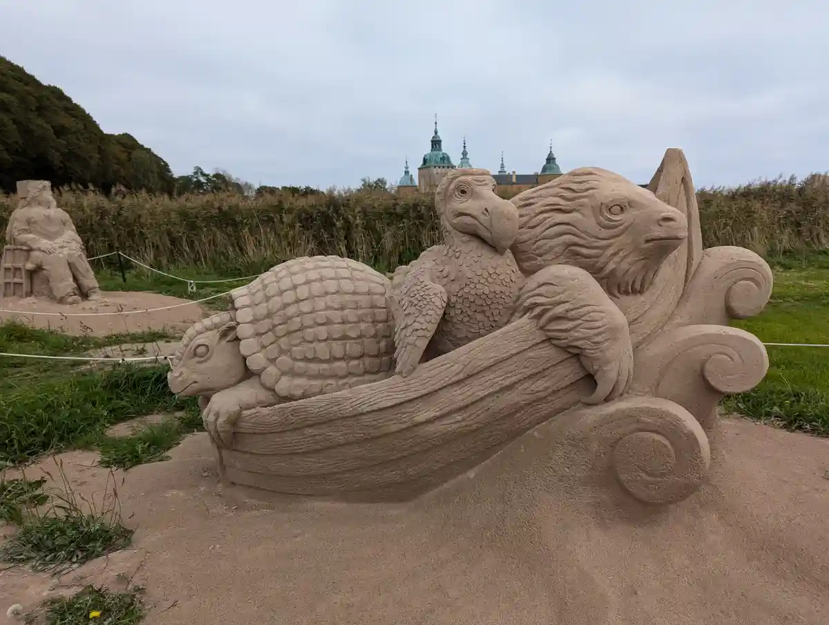 A detailed sand sculpture depicts a fantastical creature resembling a bird with a reptilian head and a turtle shell, sitting…