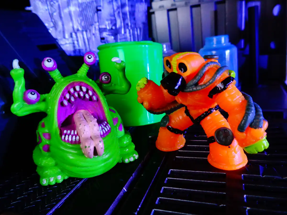 A green, multi-eyed toy monster with an open mouth full of teeth and a long tongue faces off against an orange, armored toy…