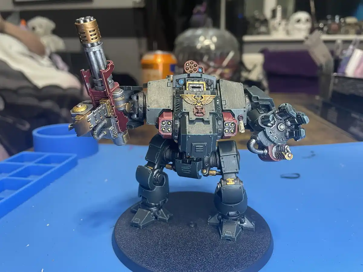 A heavily armored, custom-painted tabletop robot model stands on a blue work surface, holding a large gun in one hand and a…