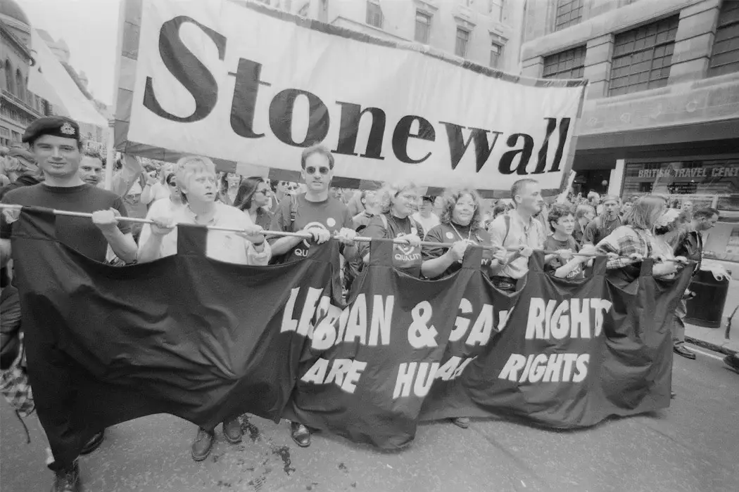 A black-and-white photo of a historical LGBTQ+ rights protest, where demonstrators carry a large banner reading 'Stonewall'…