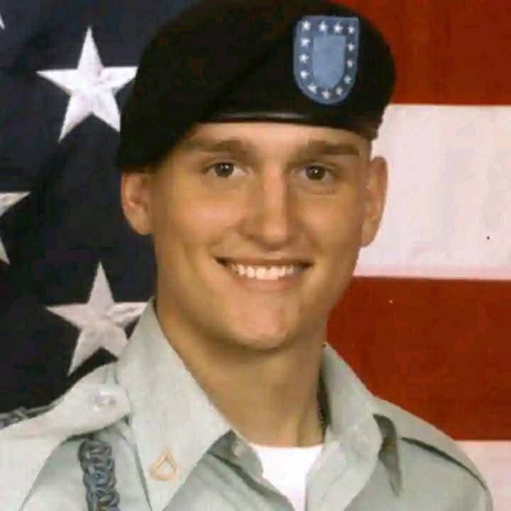 A smiling young man in a U.S. Army uniform with a black beret featuring a blue unit patch, standing in front of an American…