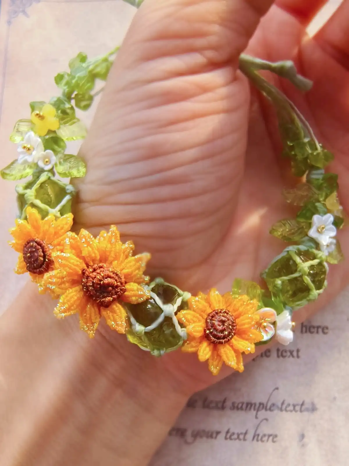 A hand holds a delicate, handmade beaded bracelet featuring intricate sunflowers and green leaves made from small, colorful…