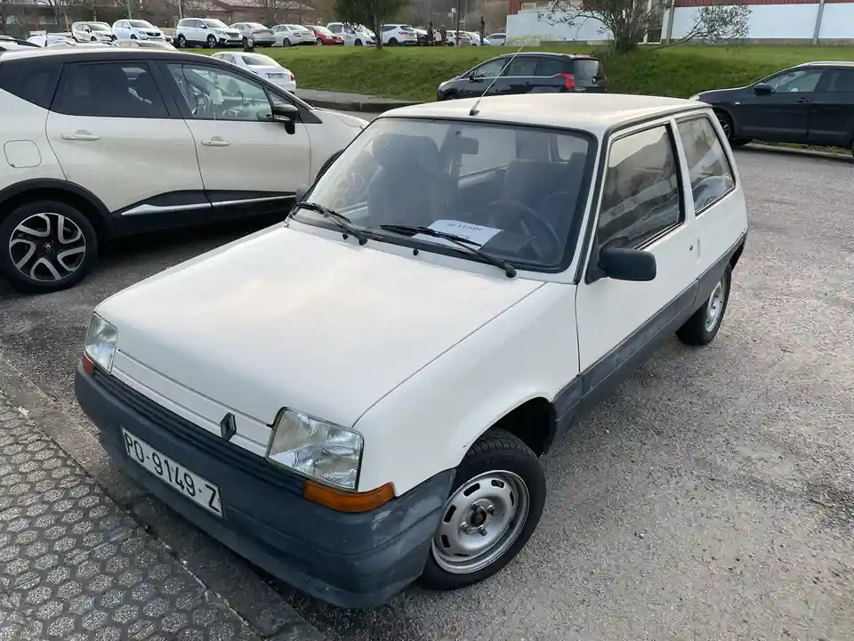 A white, boxy, older model Renault 5 parked in a lot with modern cars nearby, has a 'SE VENDE' (For Sale) sign on the…