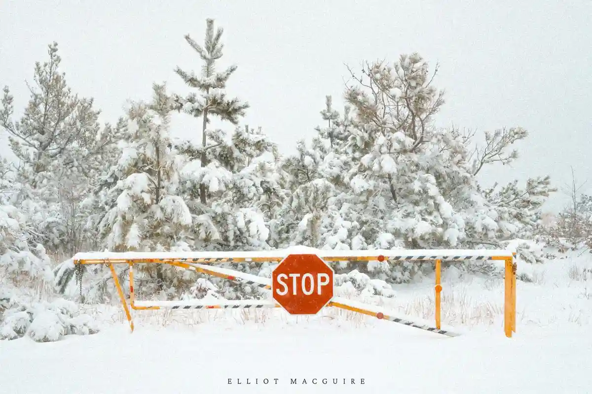 A snowy outdoor scene features a broken, snow-covered barrier gate with a red octagonal stop sign attached. The gate is…