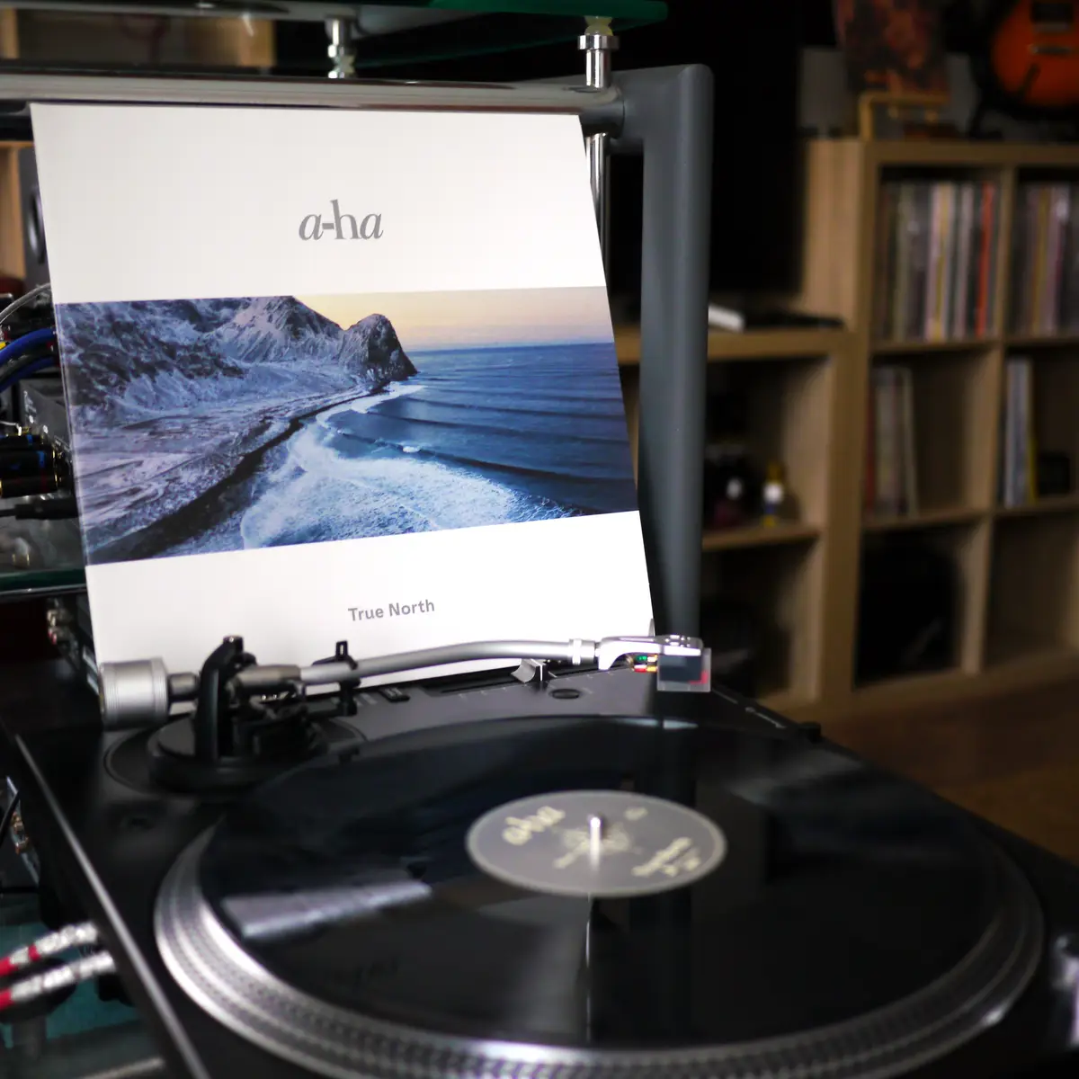 A close-up shot of a turntable with the album cover for 'True North' by a-ha standing upright behind it. The cover features…