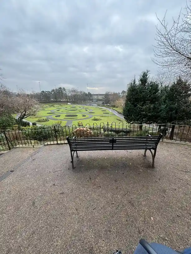 A photo shows an empty black park bench facing a large, ornate garden with circular patterns, under an overcast sky. The…