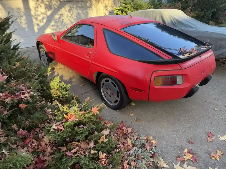 A red Porsche 924 parked in a driveway surrounded by fallen autumn leaves on bushes and pavement. A second car is covered by…