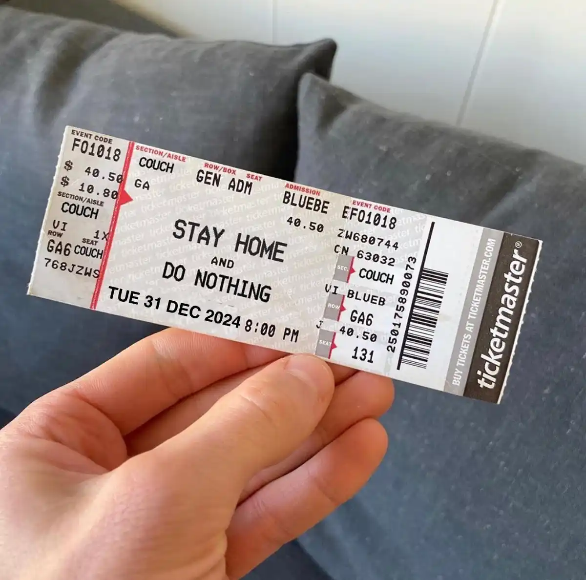 A hand holds a Ticketmaster event ticket that has been humorously altered to read 'STAY HOME AND DO NOTHING' instead of…