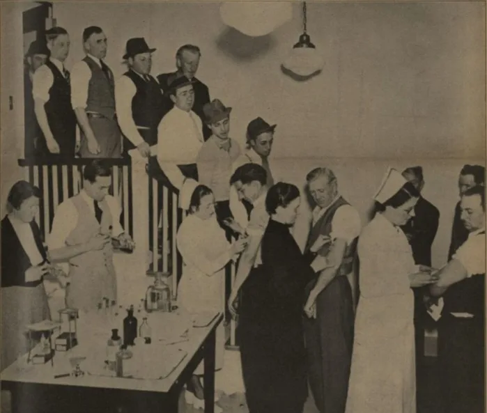 A black-and-white historical photo shows a group of men and women in early 20th-century clothing, some in nurse uniforms…