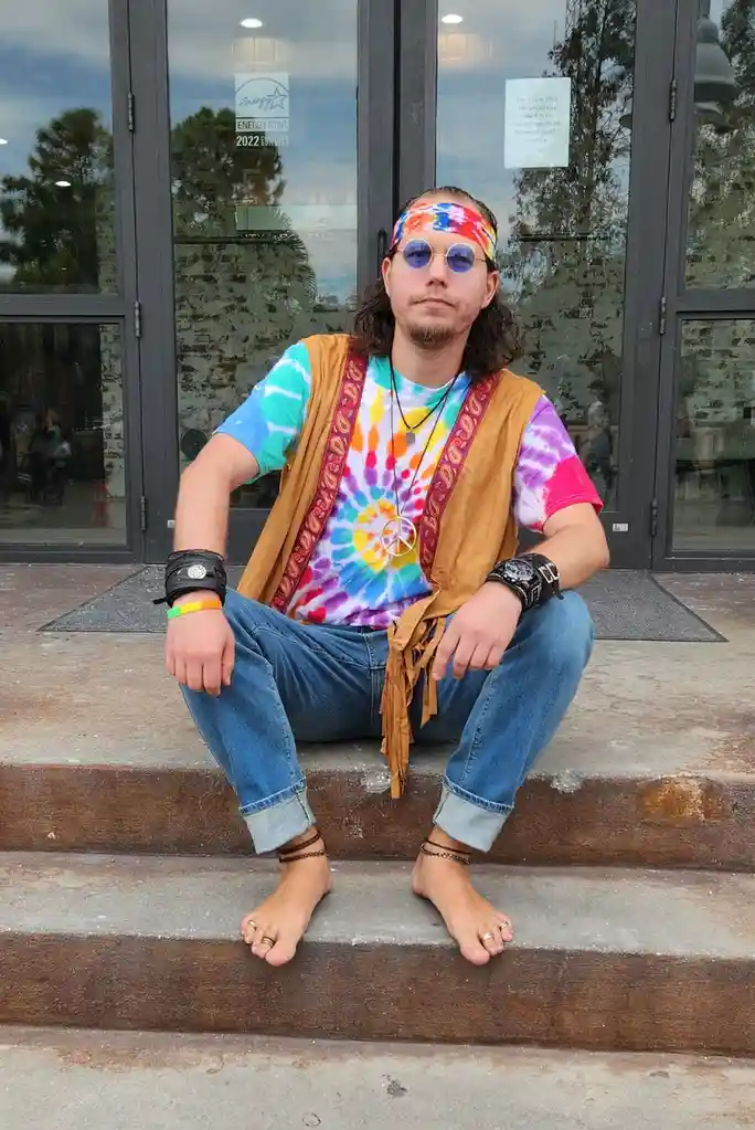 A man with long hair, wearing a tie-dye shirt, a suede vest with fringe, a peace sign necklace, round sunglasses, and a…
