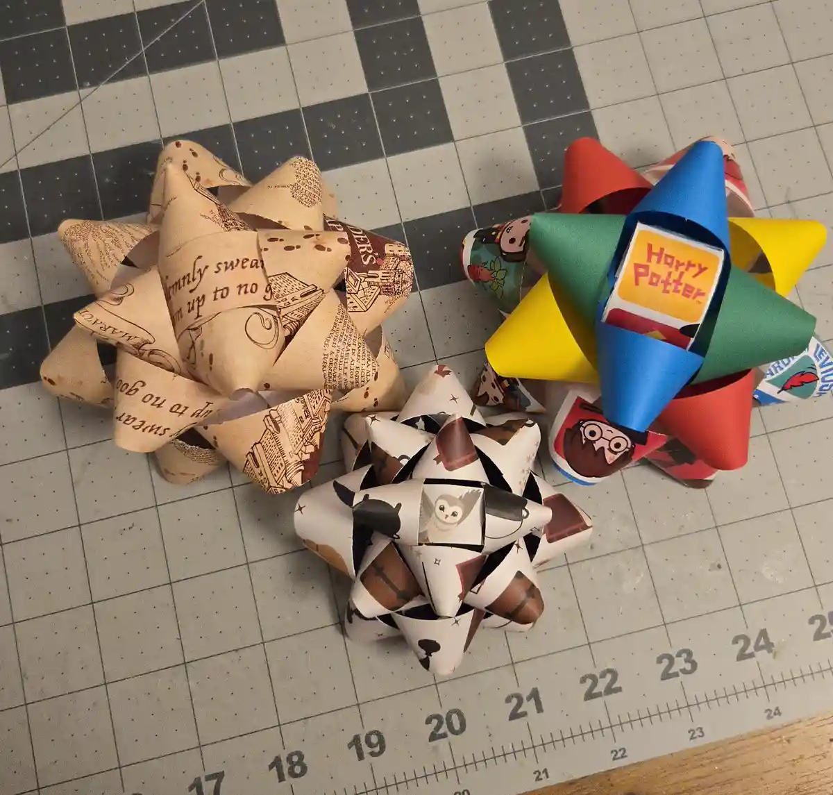 Three handmade paper bows sit on a gridded cutting mat; one is beige with Harry Potter text, one is colorful with character…