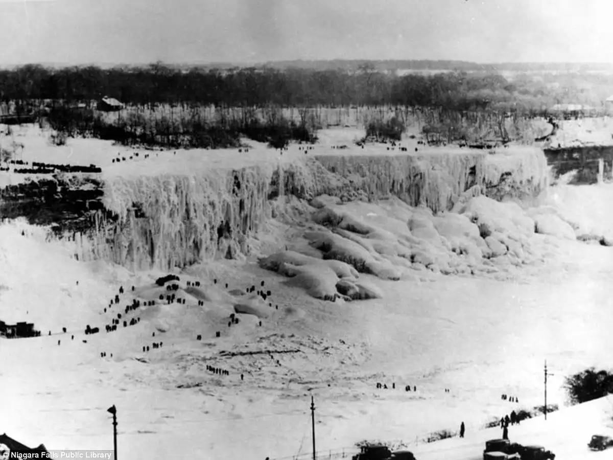 A black-and-white historical photograph shows Niagara Falls completely frozen over, with massive icicles hanging from the…