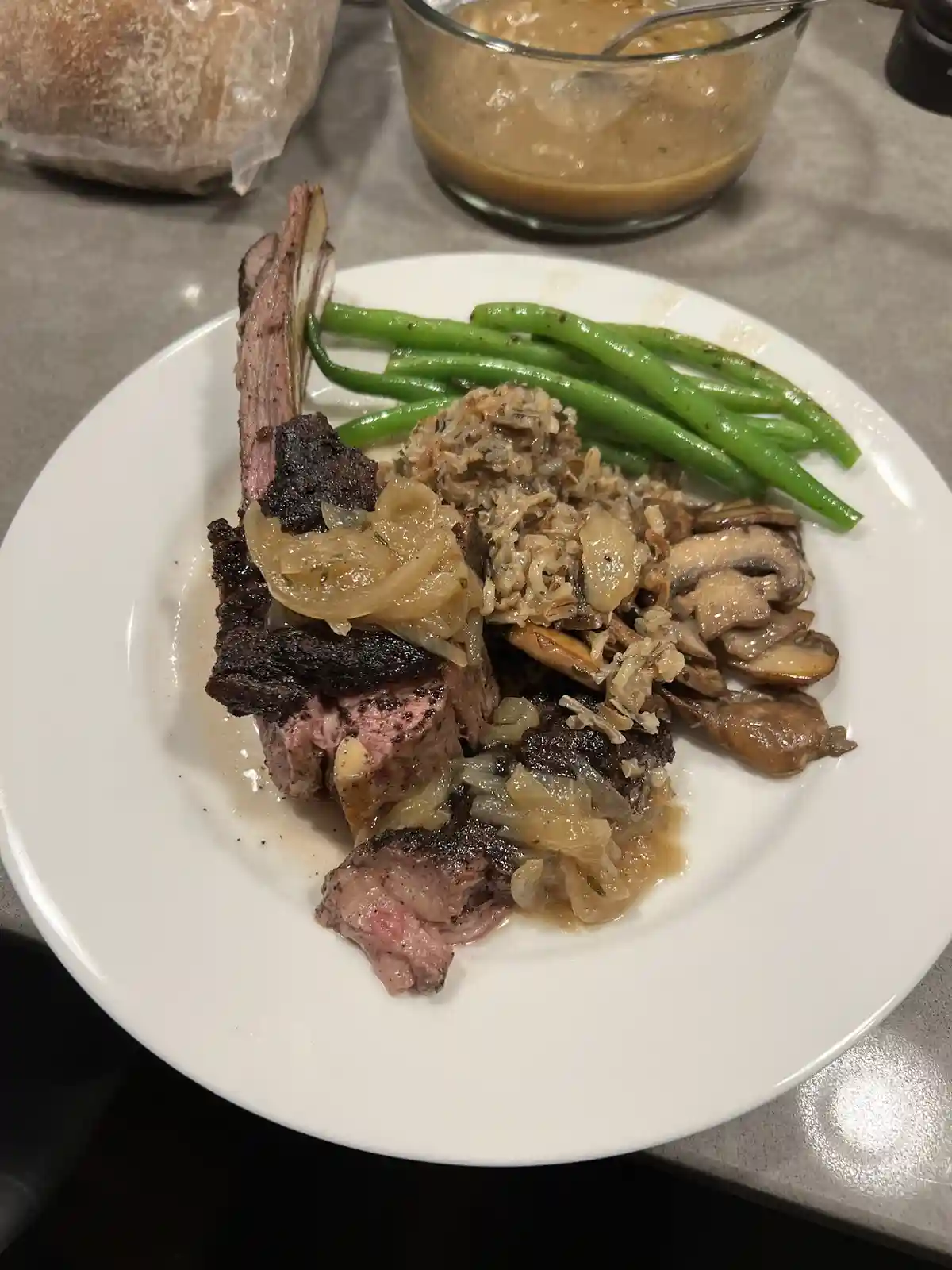 A white plate holds a cooked lamb chop with a bone, topped with caramelized onions, alongside green beans, wild rice with…