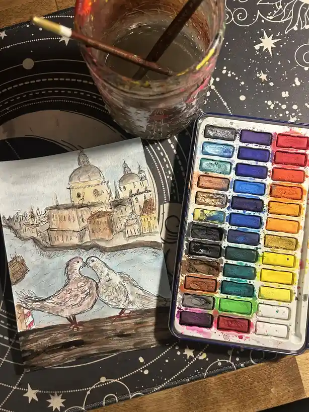 A watercolor painting of two pigeons kissing in front of domed buildings sits next to a heavily used watercolor palette and…