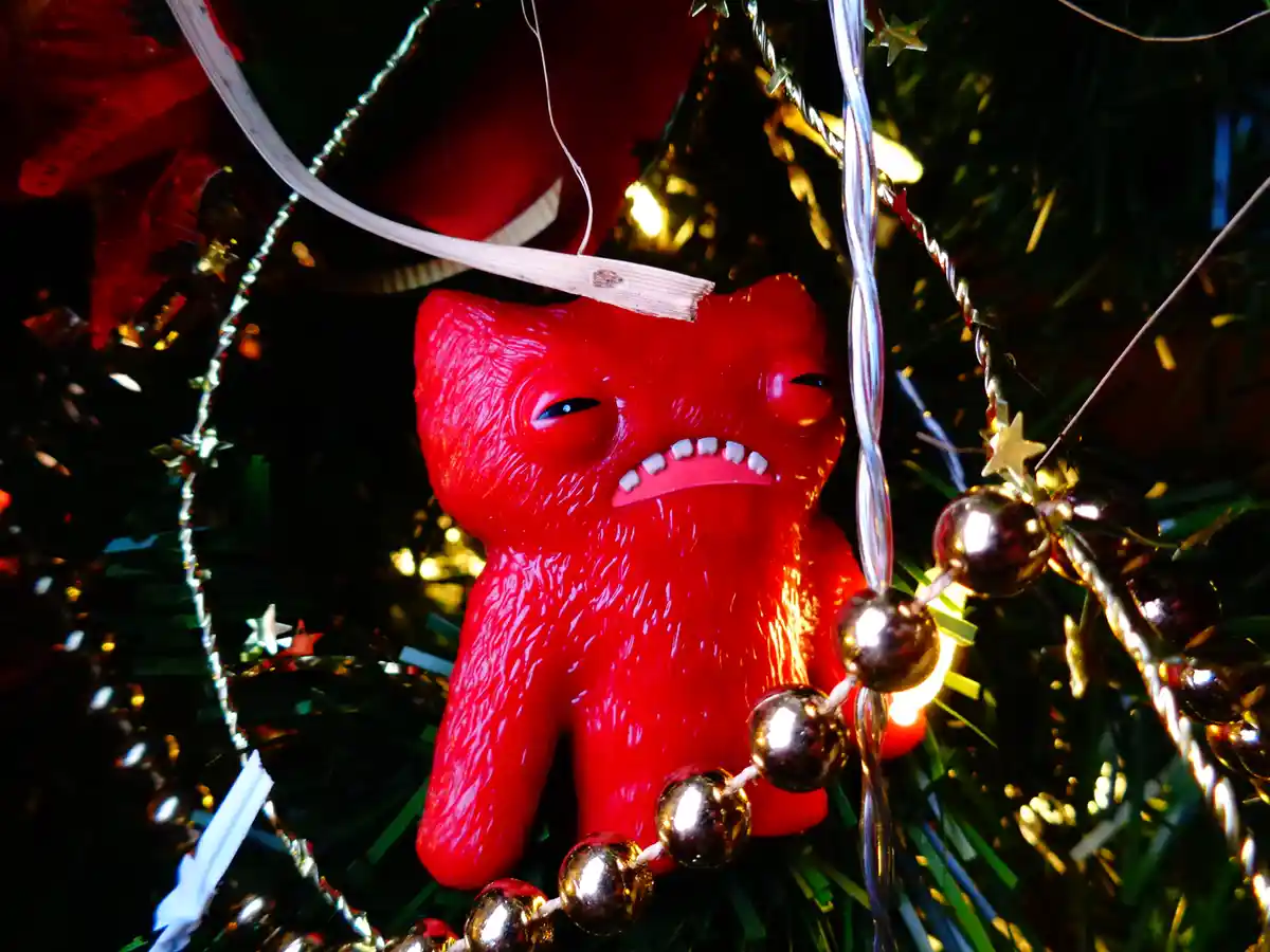 A red, textured plastic figurine resembling a grumpy, fanged demon or monster is hanging on a Christmas tree, surrounded by…