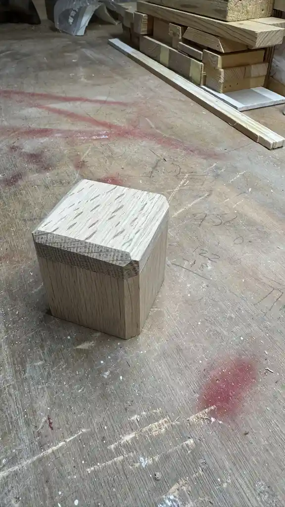 A small, handmade wooden box sits on a worn, scratched workshop floor with red spray paint marks and scattered wood…