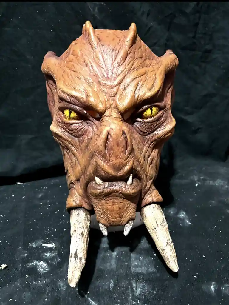 A close-up photo shows a detailed, brown, textured monster mask with glowing yellow eyes, prominent tusks, and a fierce…