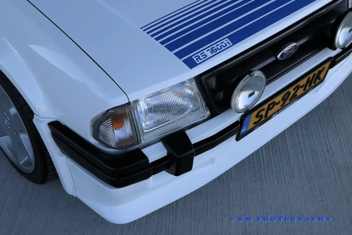 A close-up of the front of a white Ford RS 1600i, showing its blue racing stripes, headlights, grille with the Ford logo…