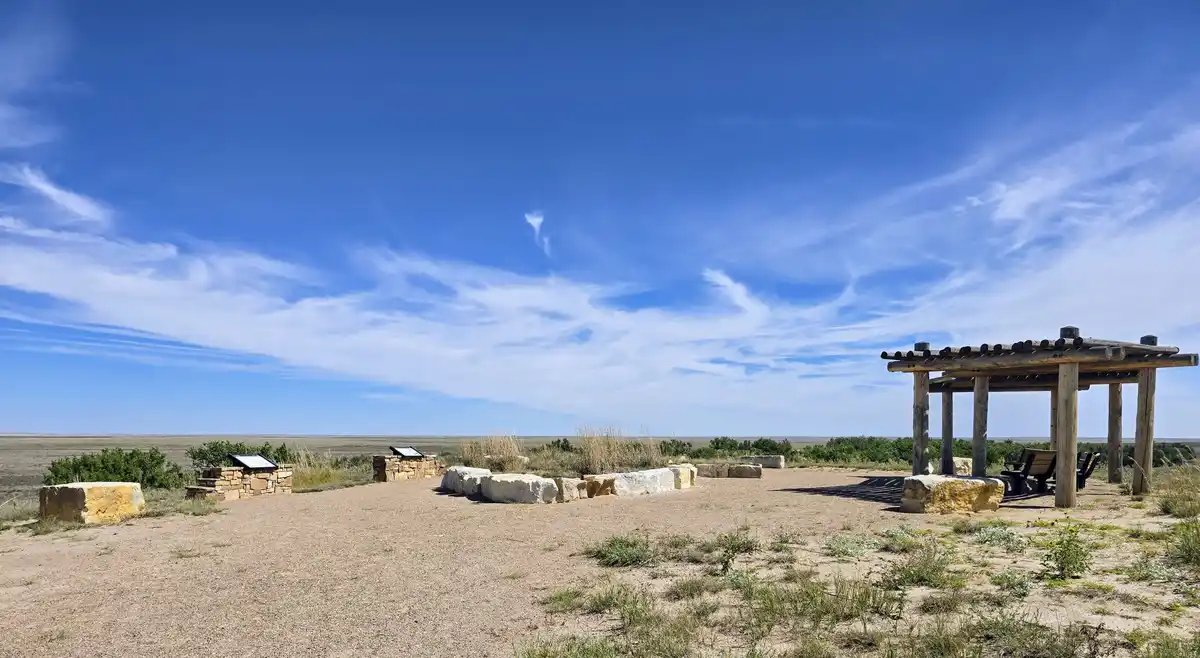A wide, sunny desert overlook with a wooden pergola, stone benches, and informational plaques under a vast blue sky with…