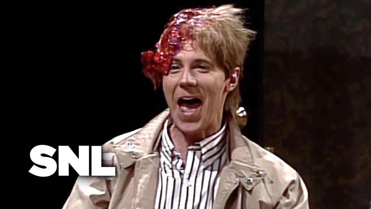 A man with spiky blonde hair has a large, gory red wound on his forehead, wearing a beige trench coat over a striped shirt…
