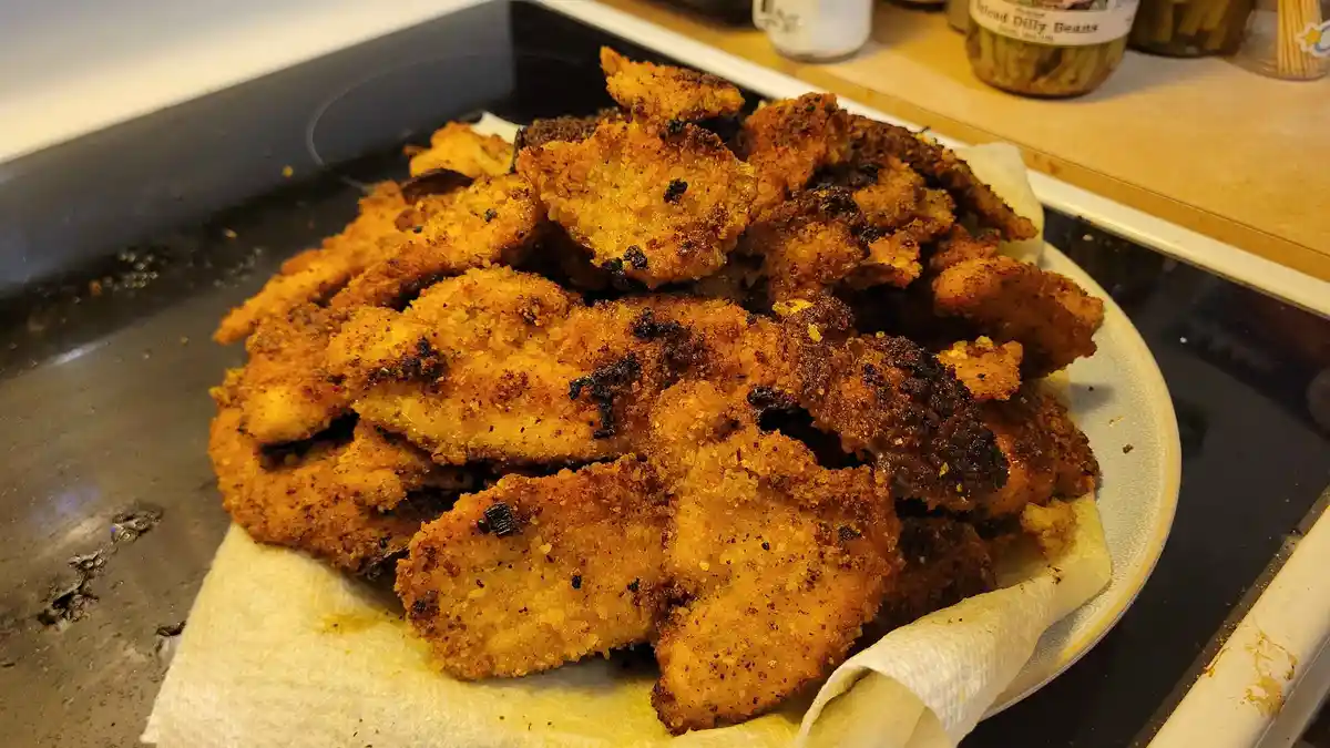 A plate of golden-brown, breaded fried chicken pieces sits on a paper towel atop a black stovetop, with some pieces showing…