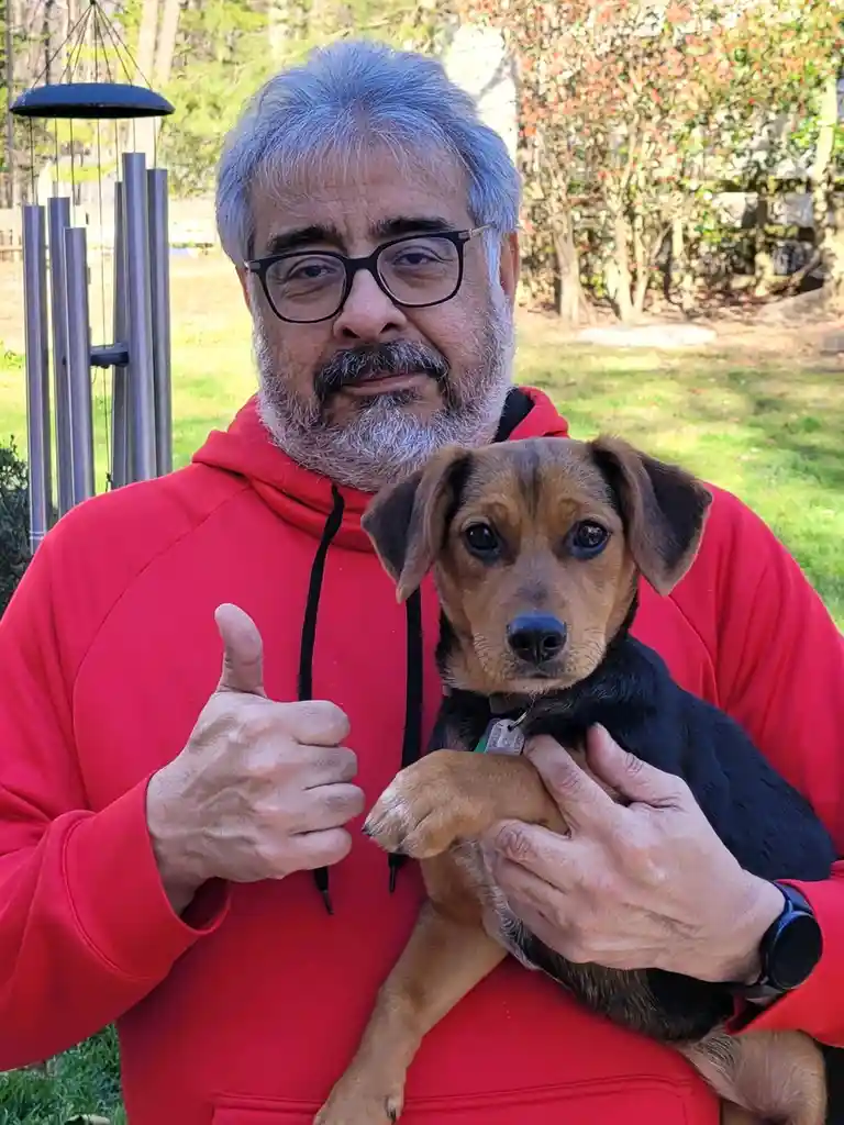 A man with gray hair and glasses, wearing a red hoodie, holds a small brown and black puppy while giving a thumbs-up. The…