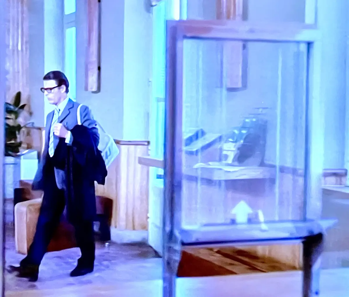 A man in a suit and glasses walks past a glass display case in a room with blue lighting. He carries a coat over his arm and…