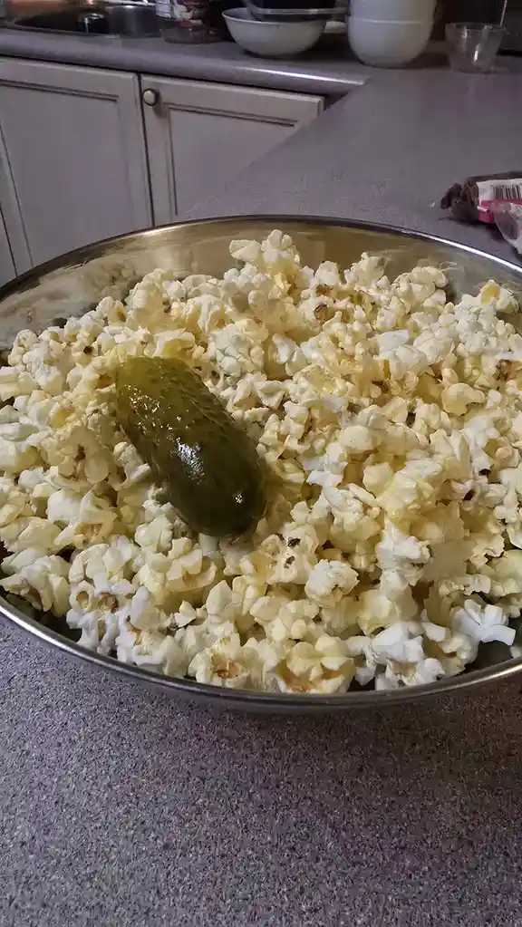 A large metal bowl filled with popcorn sits on a kitchen counter, with a single pickle resting prominently in the center…